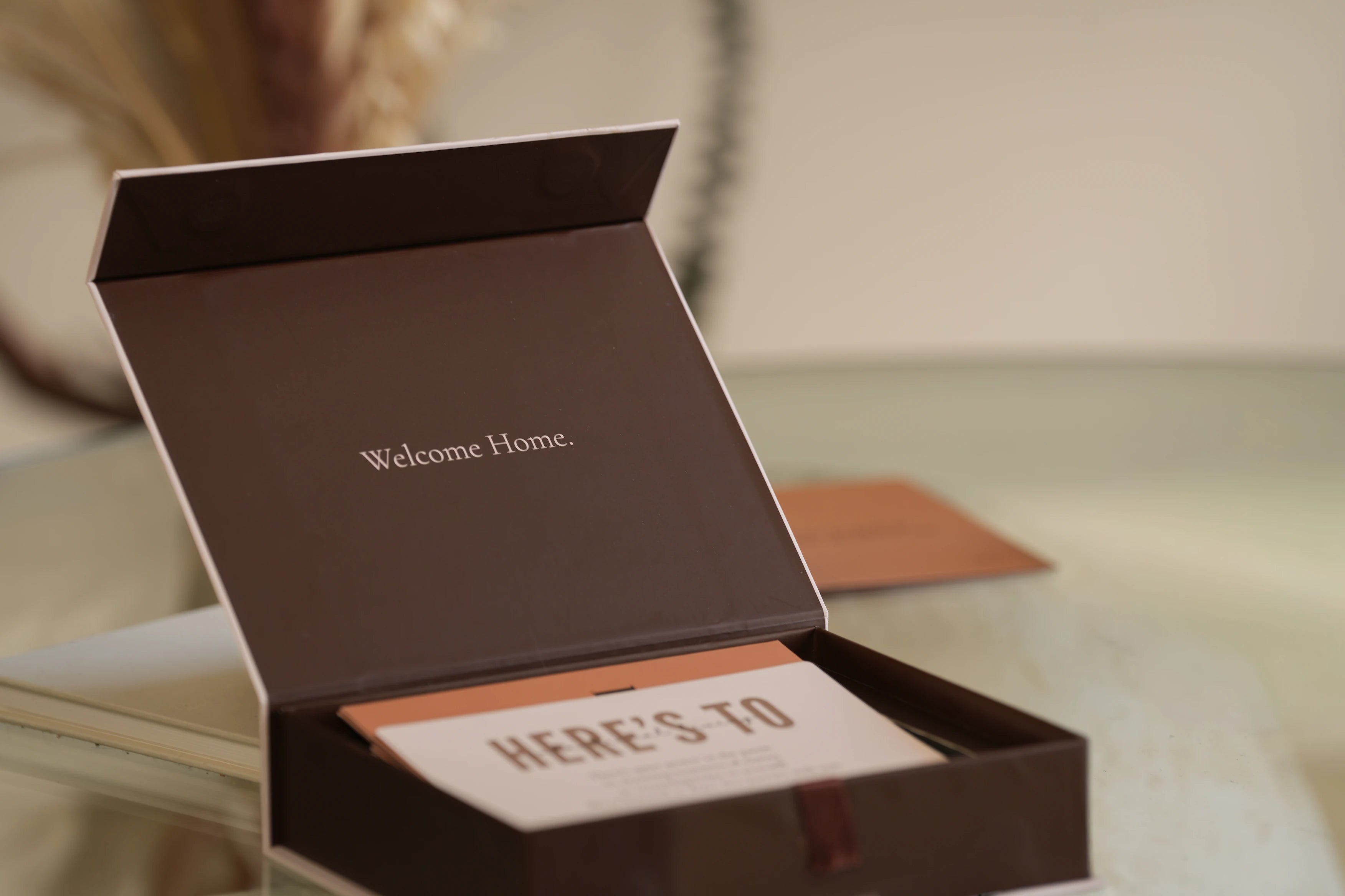Open brown box with 'Welcome Home' and 'Here's To' text on a reflective surface.