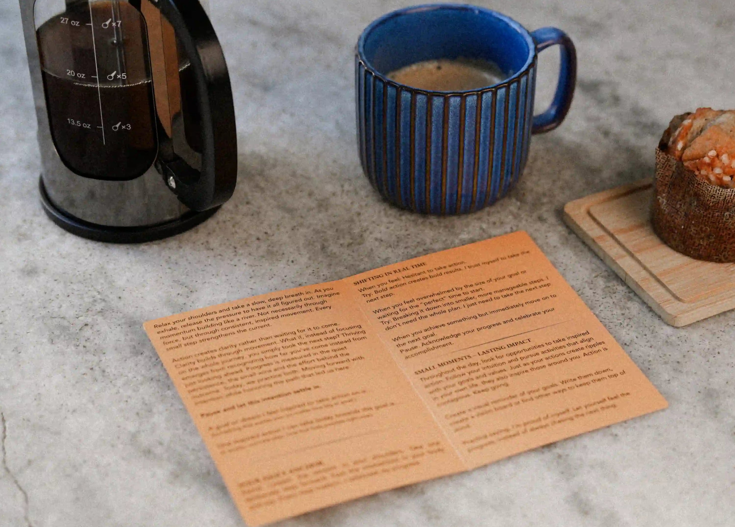 Coffee maker, blue mug with coffee, muffin, and a open blink card on a concrete surface